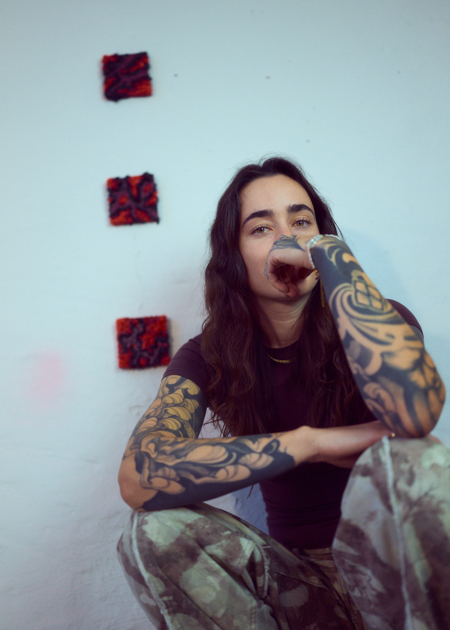 Lutza crouching in front of a white wall hung with small red and black textile works, tattooed arms resting on one knee